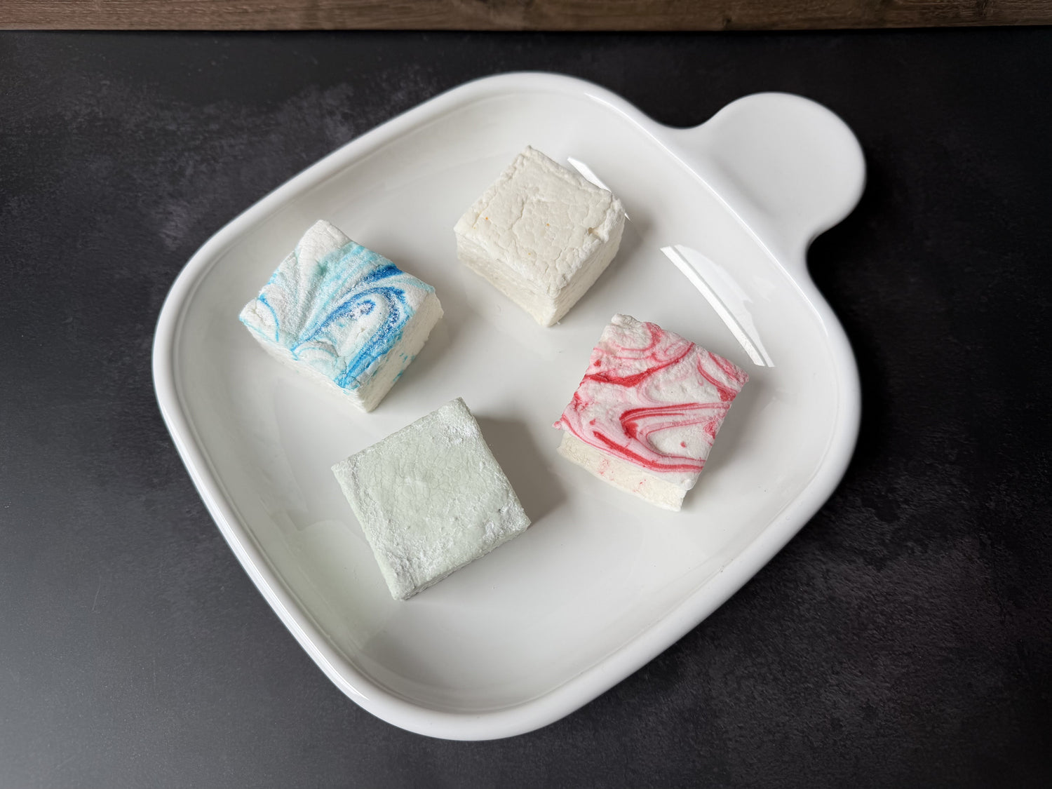Four handcrafted marshmallows representing the Customer Favorites collection, including Peppermint, Cinnamon, Key Lime, and Salted Caramel, arranged on a white ceramic plate.