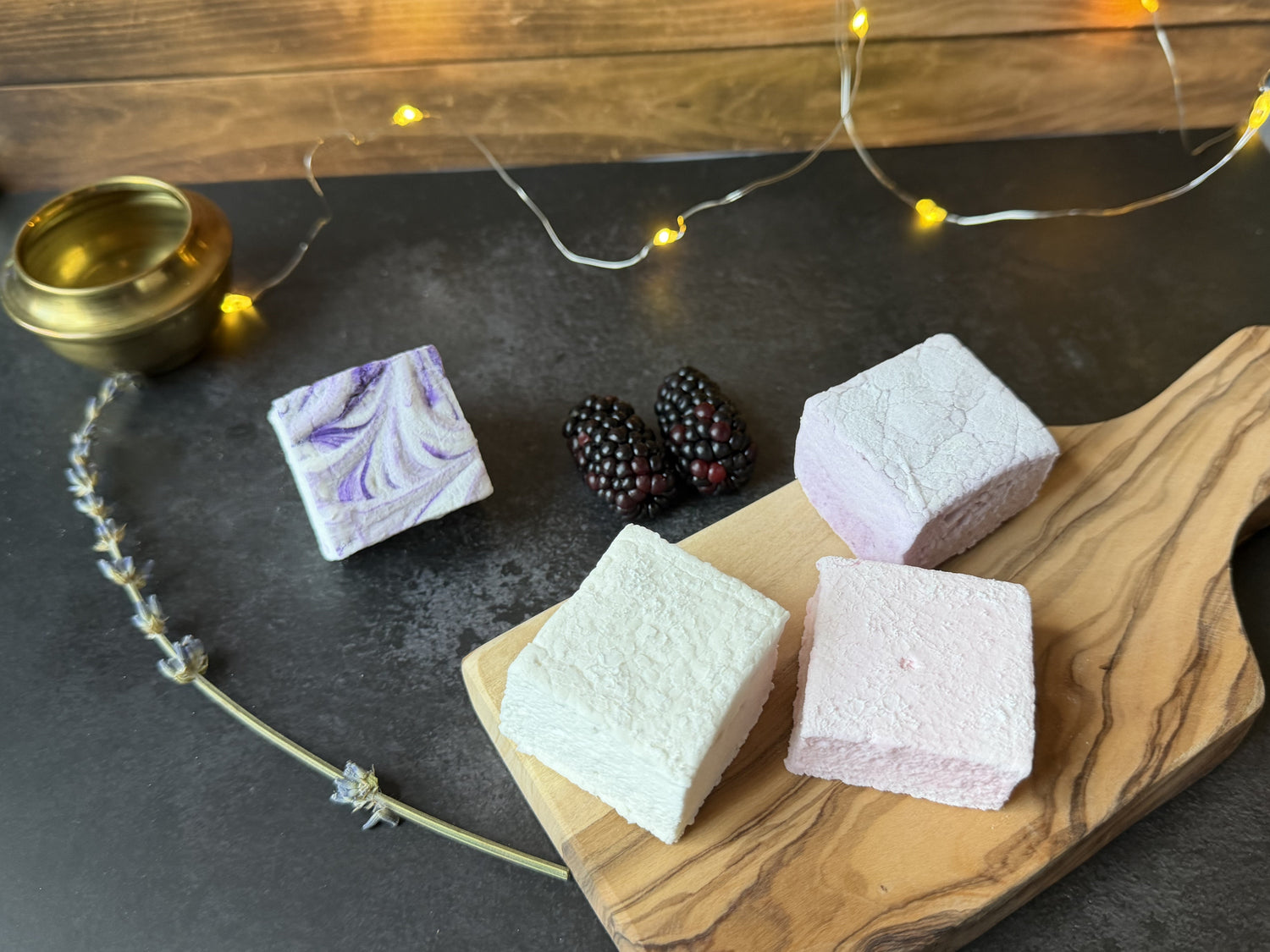 Handcrafted marshmallows from the Moonlight Magic Collection, including blackberry, lavender, vanilla, and chai flavors, styled with fairy lights on a dark surface.