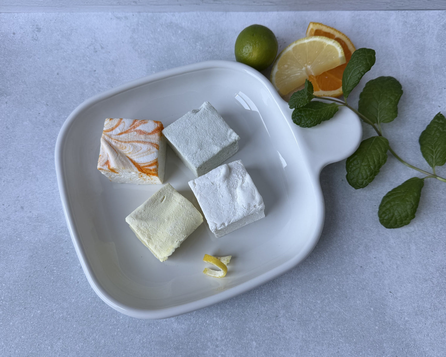Handcrafted citrus-inspired marshmallows, including lemon, orange cream, and key lime, arranged with fresh citrus on a white ceramic plate.