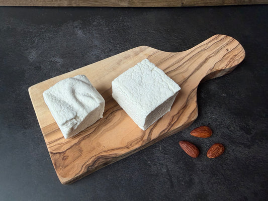 Two handcrafted Almond marshmallows on an olive wood serving board with whole almonds on a dark stone background, highlighting their soft texture and handmade appearance.
