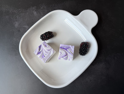 Handcrafted Blackberry marshmallows with purple swirls on a white ceramic plate with fresh blackberries on a dark stone surface. Two marshmallows showcase their soft, fluffy texture and handcrafted swirl pattern.