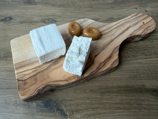 Handcrafted Butterscotch marshmallows on an olive wood board with butterscotch candies on a warm wood tabletop. One marshmallow is whole and one is torn open to reveal its soft, fluffy texture.