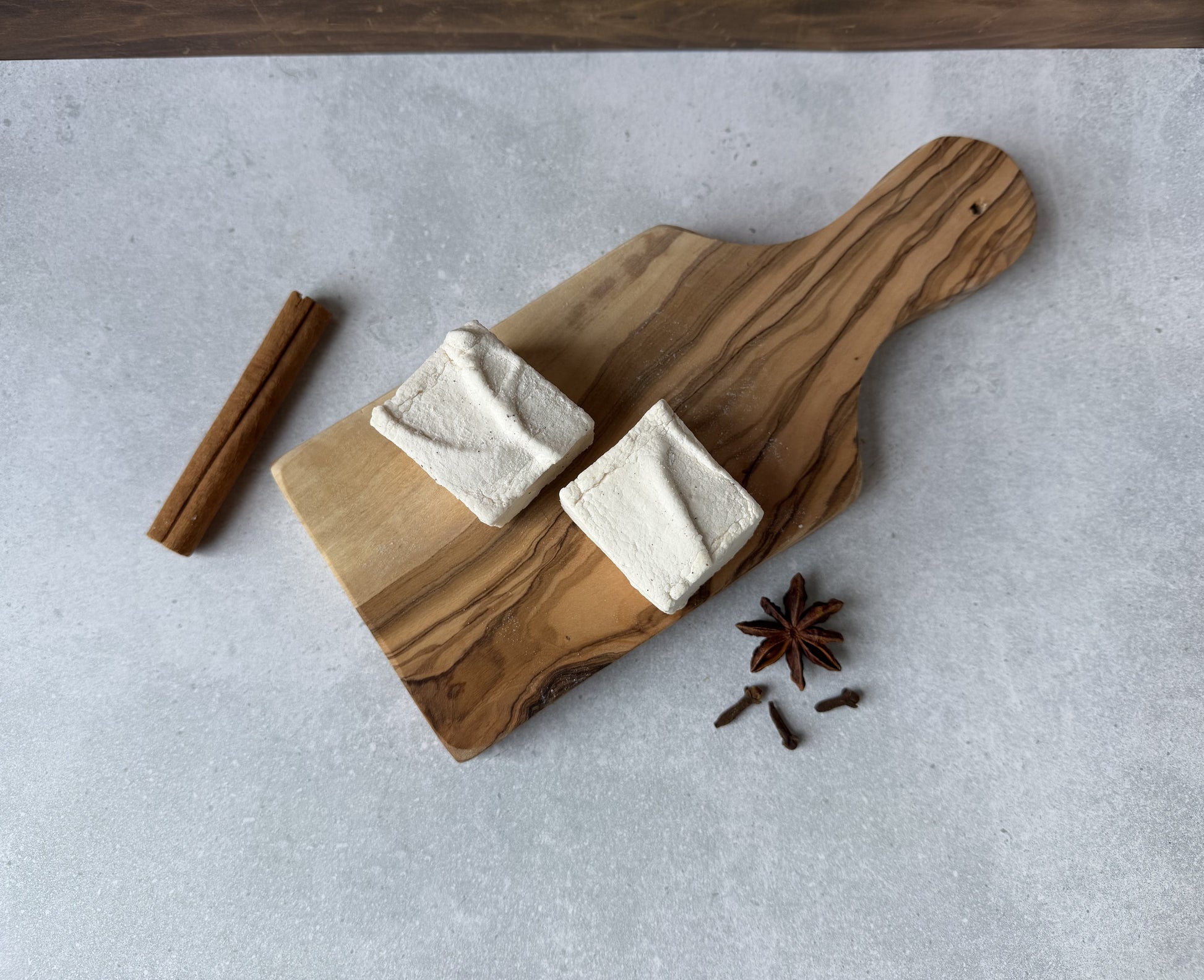 Handcrafted Chai marshmallows on an olive wood serving board with cinnamon sticks, star anise, and whole cloves on a light gray stone surface. Two marshmallows showcase their soft, fluffy texture and warm chai-inspired flavor.