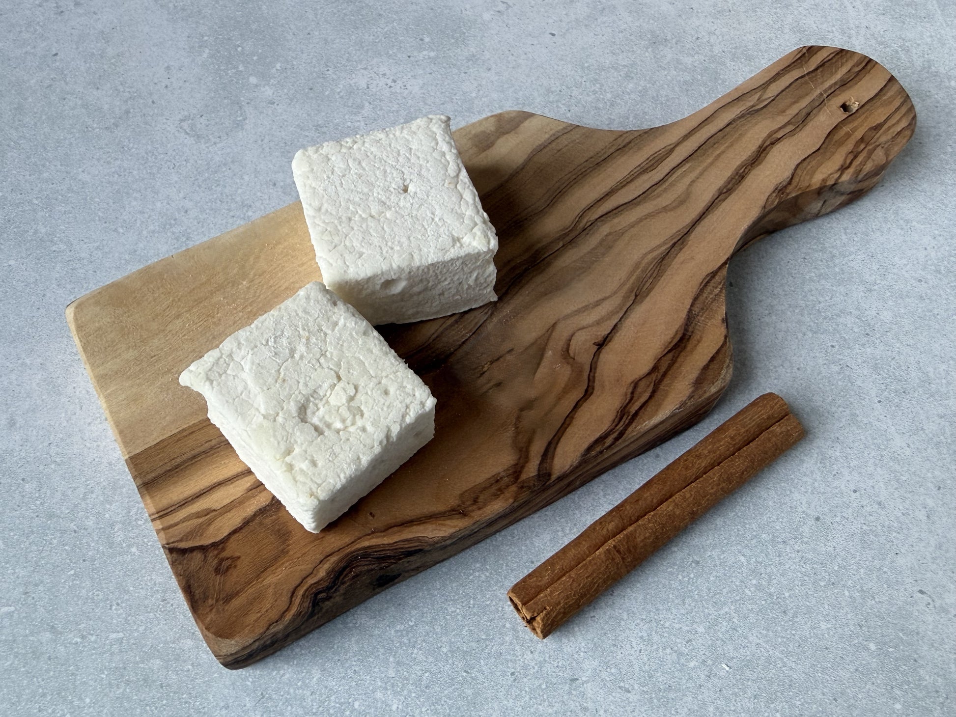 Handcrafted Cinnamon marshmallows on an olive wood serving board with cinnamon sticks on a light gray stone surface. Two marshmallows showcase their soft, fluffy texture and handcrafted appearance.