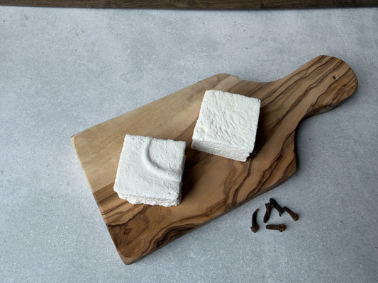 Two handcrafted Clove marshmallows on an olive wood serving board with whole cloves on a light gray stone background, highlighting their soft texture and handmade appearance.