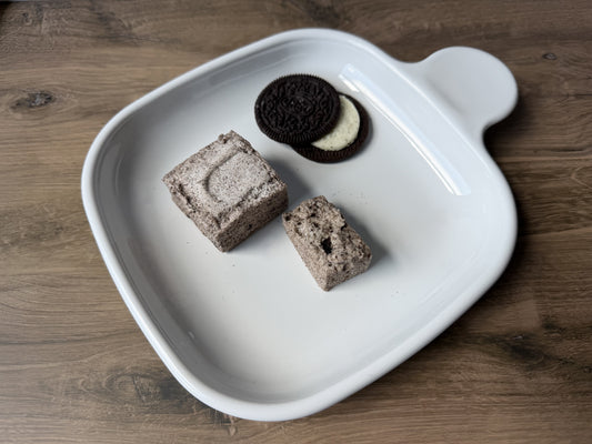 Handcrafted Cookies & Cream marshmallows on a white ceramic plate with a twisted Oreo cookie. One marshmallow is whole and one is cut open to show chocolate cookie pieces inside.