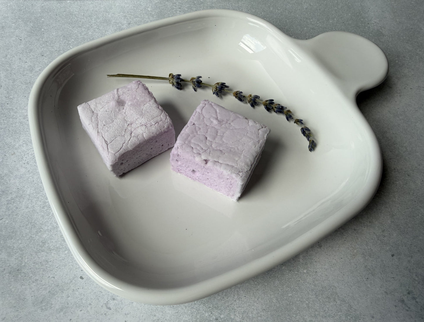 Handcrafted Lavender marshmallows on a white ceramic plate with a sprig of dried lavender. Two marshmallows showcase their soft lavender color and fluffy handcrafted texture.