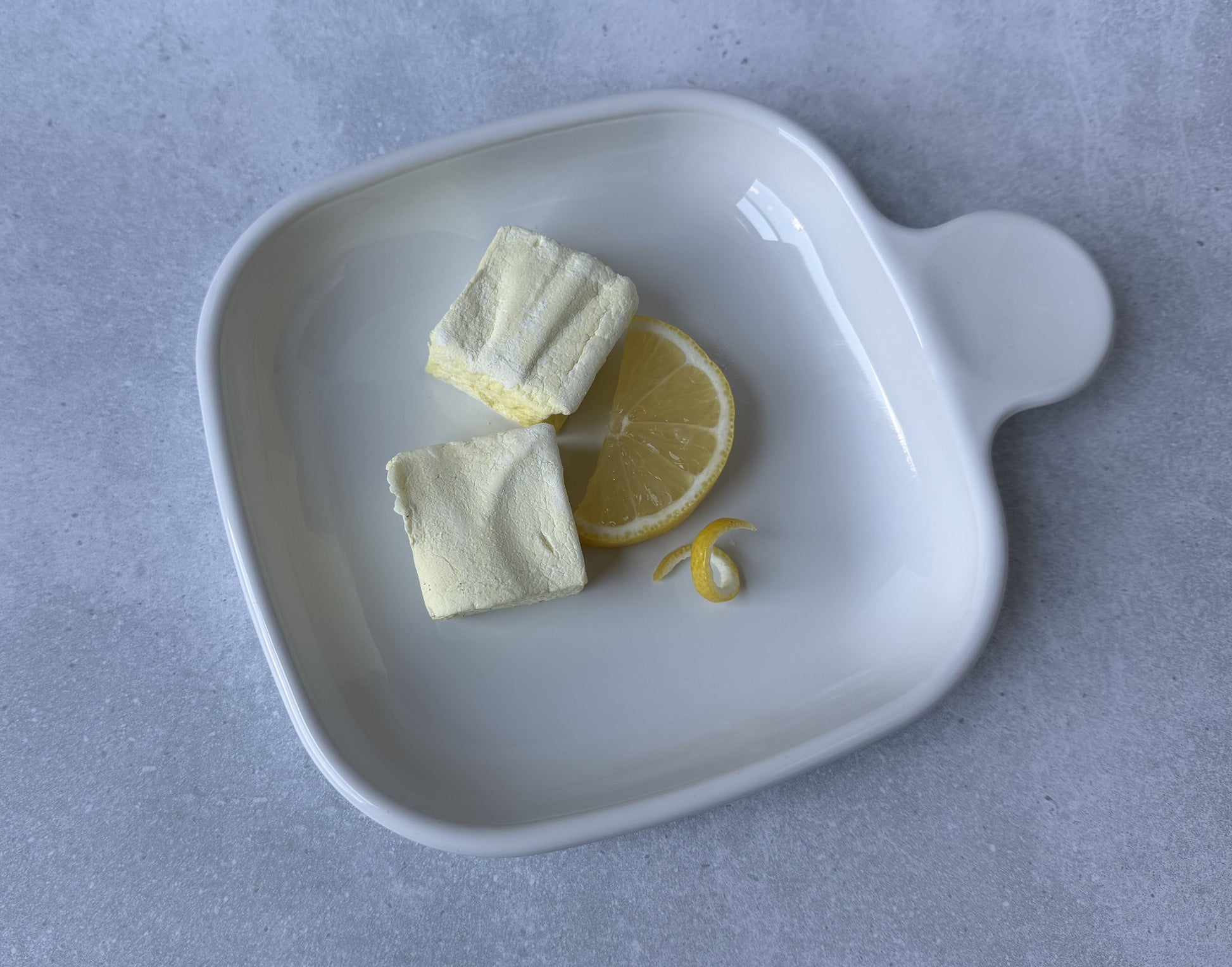 Handcrafted Lemon marshmallows on a white ceramic plate with a fresh lemon slice and lemon peel twist on a light gray stone surface. Two marshmallows showcase their soft, fluffy texture and delicate yellow color.