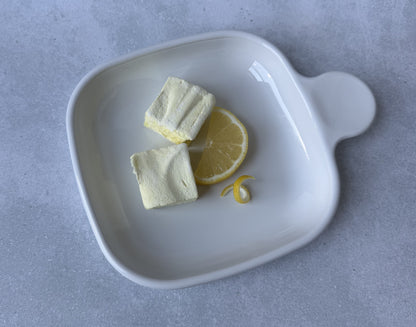 Handcrafted Lemon marshmallows on a white ceramic plate with a fresh lemon slice and lemon peel twist on a light gray stone surface. Two marshmallows showcase their soft, fluffy texture and delicate yellow color.