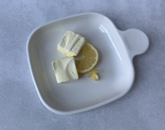 Handcrafted Lemon marshmallows on a white ceramic plate with a fresh lemon slice and lemon peel twist on a light gray stone surface. Two marshmallows showcase their soft, fluffy texture and delicate yellow color.