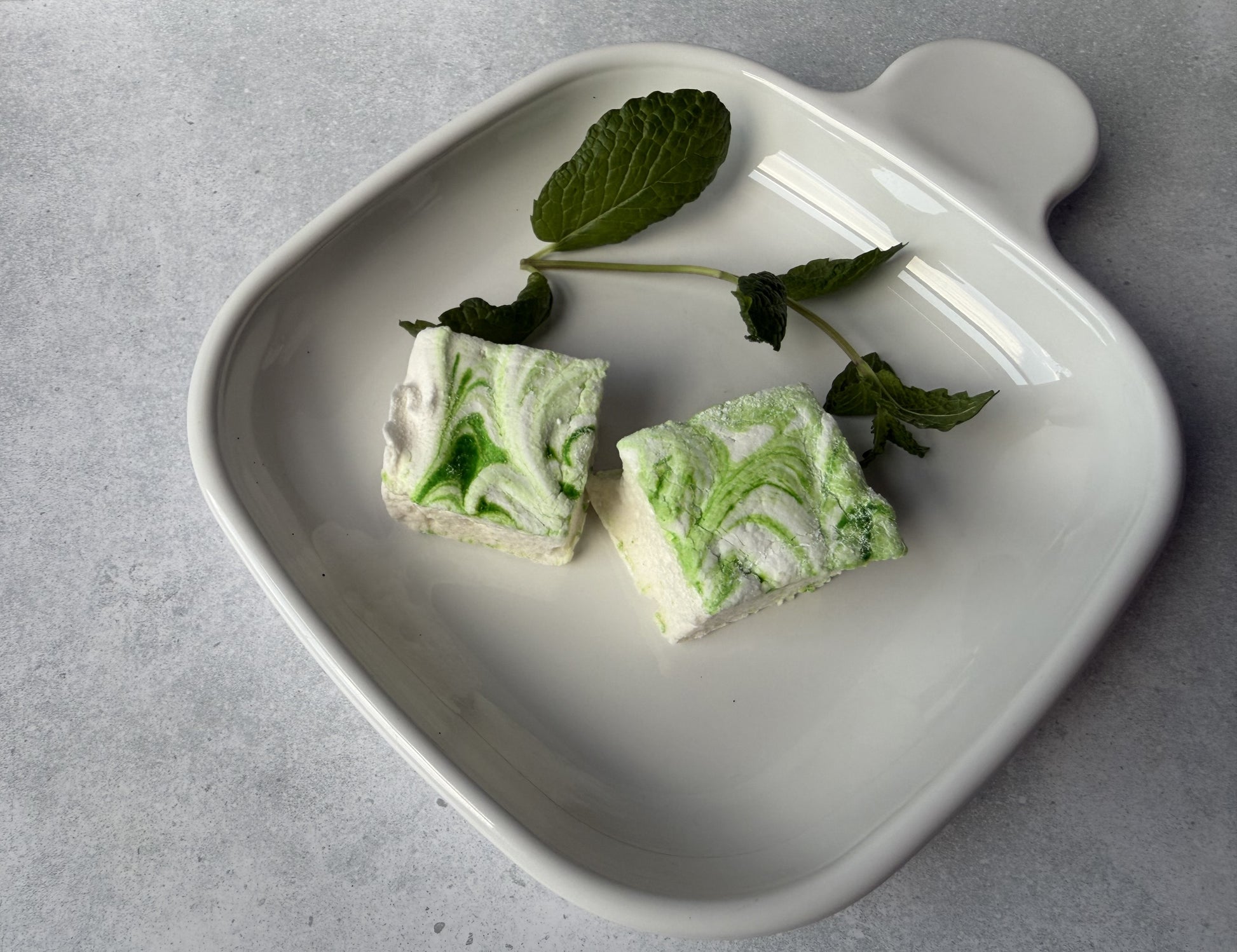 Handcrafted Mint marshmallows with green swirls on a white ceramic plate with fresh mint leaves. Two marshmallows are arranged to highlight their handcrafted design and soft texture.