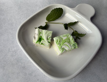 Handcrafted Mint marshmallows with green swirls on a white ceramic plate with fresh mint leaves. Two marshmallows are arranged to highlight their handcrafted design and soft texture.