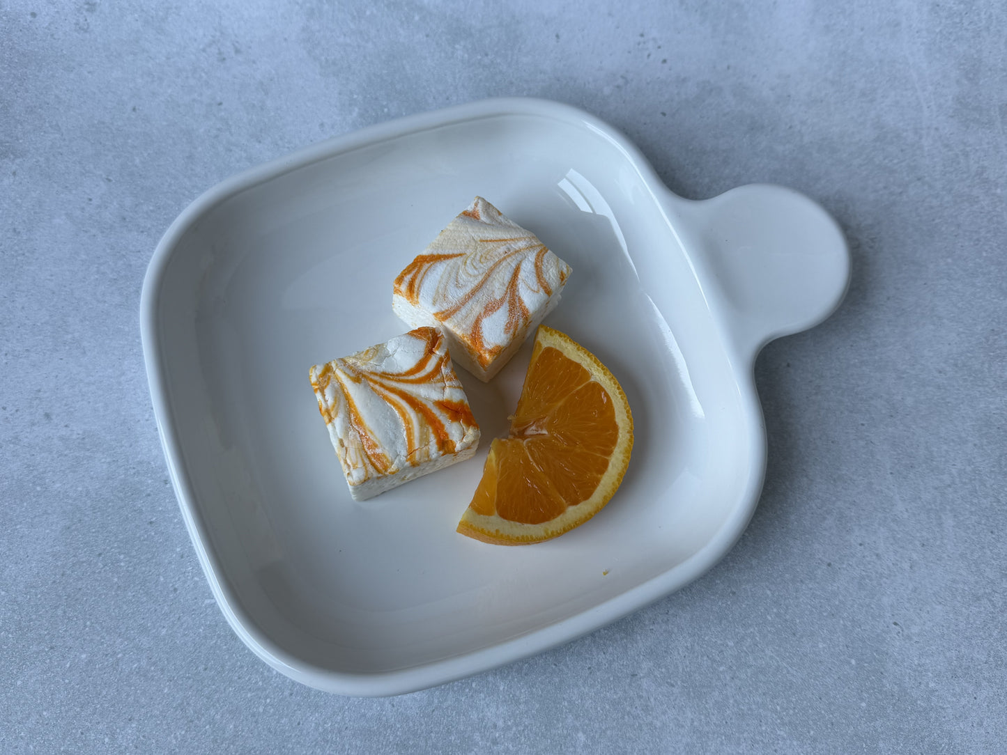 Handcrafted Orange Cream marshmallows with orange and white swirls on a white ceramic plate with a fresh orange slice. Two marshmallows showcase their soft, fluffy texture on a light gray stone background.