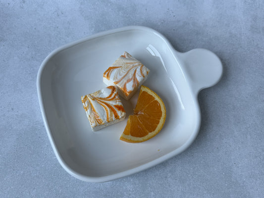 Handcrafted Orange Cream marshmallows with orange and white swirls on a white ceramic plate with a fresh orange slice. Two marshmallows showcase their soft, fluffy texture on a light gray stone background.