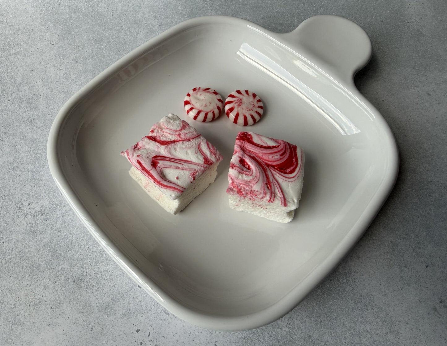 Handcrafted Peppermint marshmallows with red peppermint swirls on a white ceramic plate beside peppermint candies. The marshmallows feature a soft, fluffy texture and festive handcrafted design.