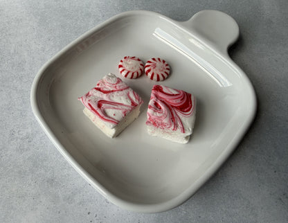 Handcrafted Peppermint marshmallows with red peppermint swirls on a white ceramic plate beside peppermint candies. The marshmallows feature a soft, fluffy texture and festive handcrafted design.