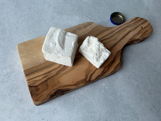 Handcrafted Root Beer Float marshmallows on an olive wood board with a vintage bottle cap on a light gray stone surface. One marshmallow is whole and one is torn open to show its fluffy interior.