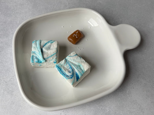 Handcrafted Salted Caramel marshmallows with blue swirls on a white ceramic plate with a caramel candy and flaky sea salt on a light gray stone surface. Two marshmallows showcase their soft, fluffy texture and handcrafted design.