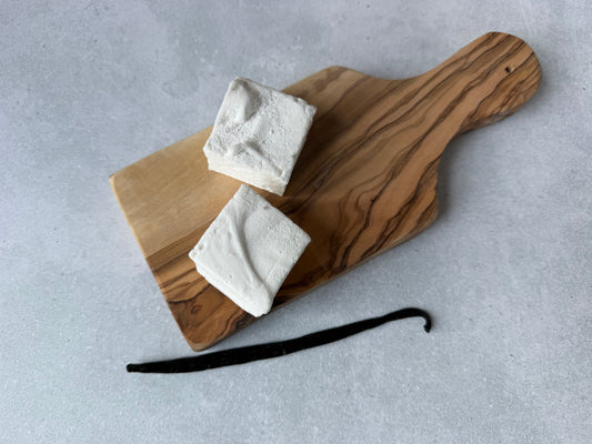 Handcrafted Vanilla marshmallows on an olive wood serving board with a whole vanilla bean on a light gray stone background. Two marshmallows highlight their soft, fluffy texture and handcrafted appearance.